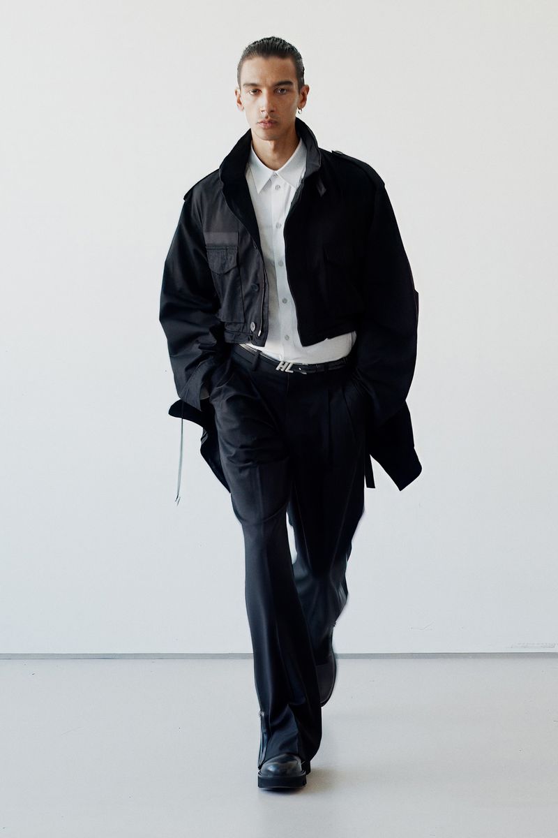 Male fashion model on white backdrop wearing black jacket, white shirt, black trousers, black shoes, and a silver belt.