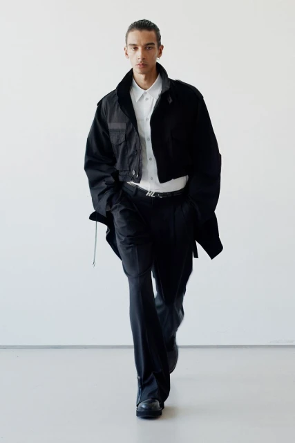 Male fashion model on white backdrop wearing black jacket, white shirt, black trousers, black shoes, and a silver belt.