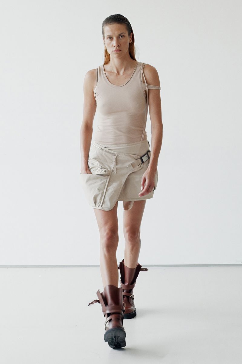 A fashionable adult woman in a neutral tank top, wrap skirt, and lug boots walks toward the camera against a white backdrop in a studio.