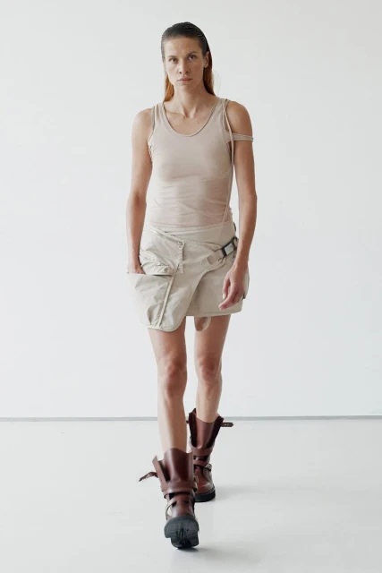 A fashionable adult woman in a neutral tank top, wrap skirt, and lug boots walks toward the camera against a white backdrop in a studio.