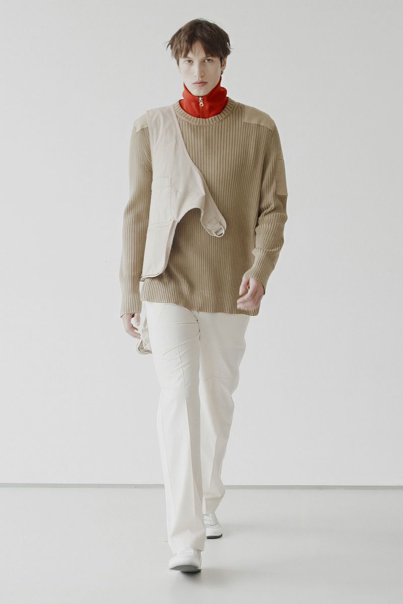 Fashionable adult male model in light brown ribbed sweater and white pants, walking on white studio background. Fashion editorial photography.