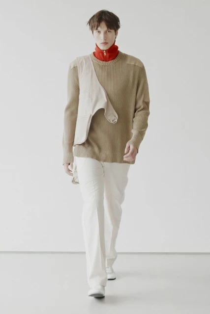 Fashionable adult male model in light brown ribbed sweater and white pants, walking on white studio background. Fashion editorial photography.