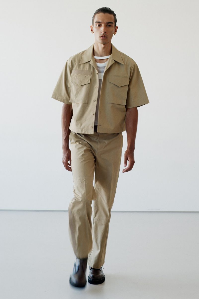 Male fashion model in tan, short-sleeved shirt and tan trousers walks in a studio. He's wearing dark shoes, looking forward, neutral facial expression.