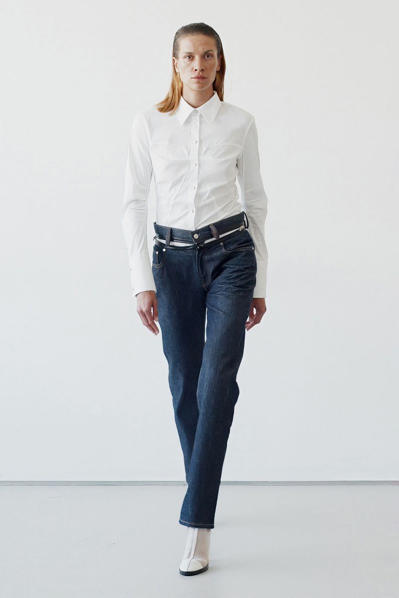 Full-length shot of a woman in a white button-down shirt and dark wash jeans against a white backdrop, captured in a fashion-forward style.