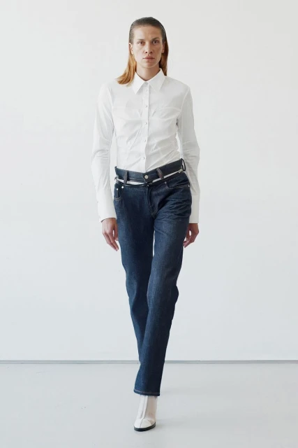 Full-length shot of a woman in a white button-down shirt and dark wash jeans against a white backdrop, captured in a fashion-forward style.