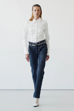 Full-length shot of a woman in a white button-down shirt and dark wash jeans against a white backdrop, captured in a fashion-forward style.
