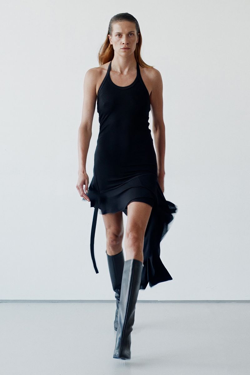 Model in a black halter dress and tall boots walking forward against a white studio background