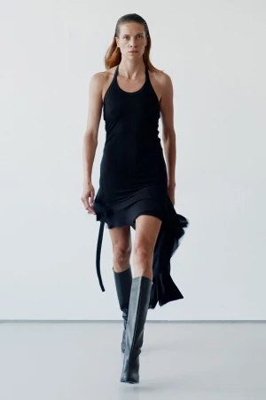 Model in a black halter dress and tall boots walking forward against a white studio background