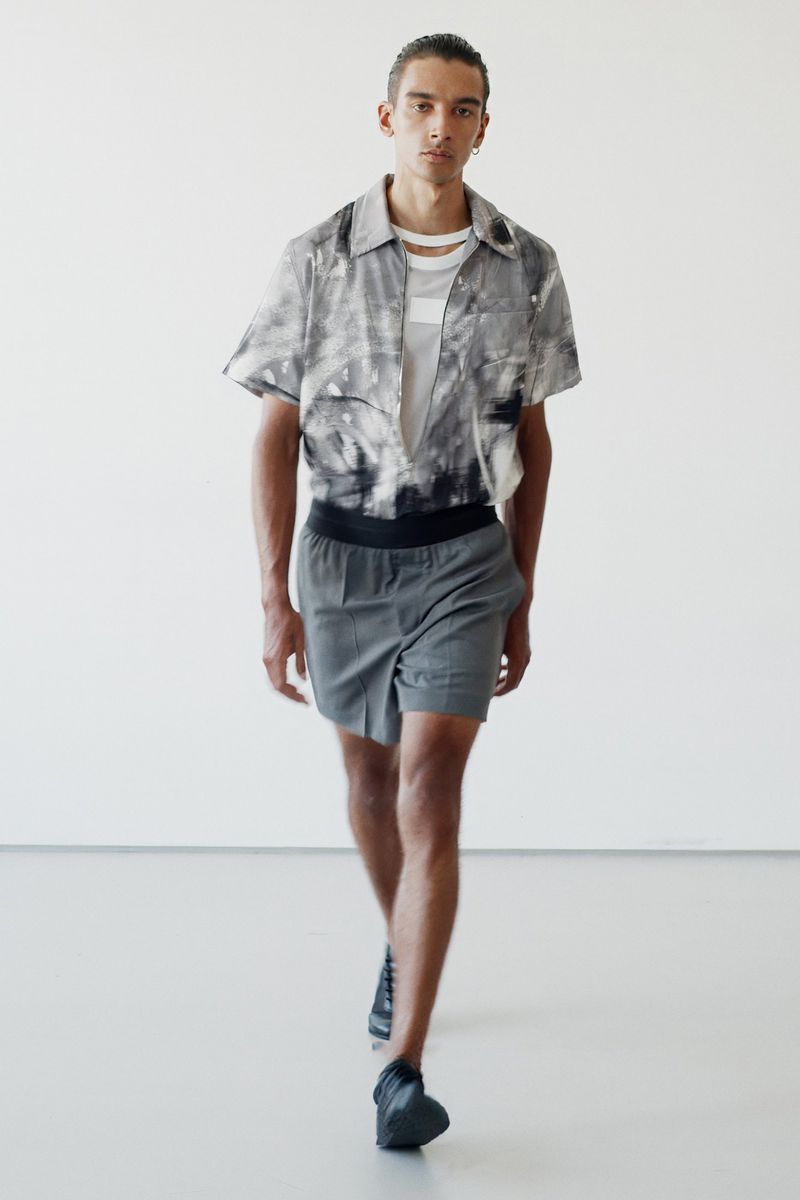Fashion shot of a male model in a patterned short-sleeved shirt and grey shorts walking in a brightly lit photo studio.