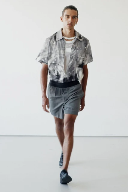 Fashion shot of a male model in a patterned short-sleeved shirt and grey shorts walking in a brightly lit photo studio.