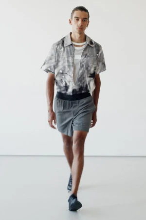 Fashion shot of a male model in a patterned short-sleeved shirt and grey shorts walking in a brightly lit photo studio.