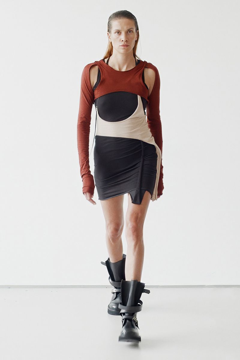 A slender fashion model strides forward confidently wearing an avant-garde black dress, rust-colored cropped top, and black boots on a white background.
