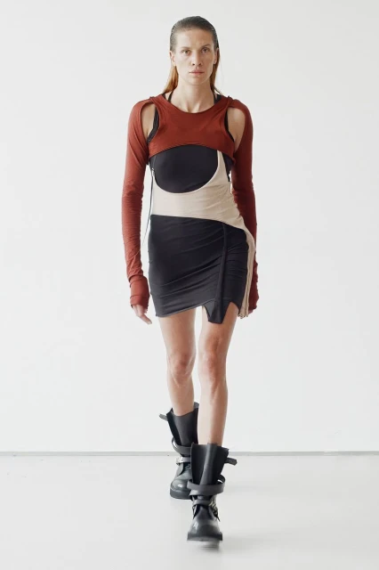 A slender fashion model strides forward confidently wearing an avant-garde black dress, rust-colored cropped top, and black boots on a white background.