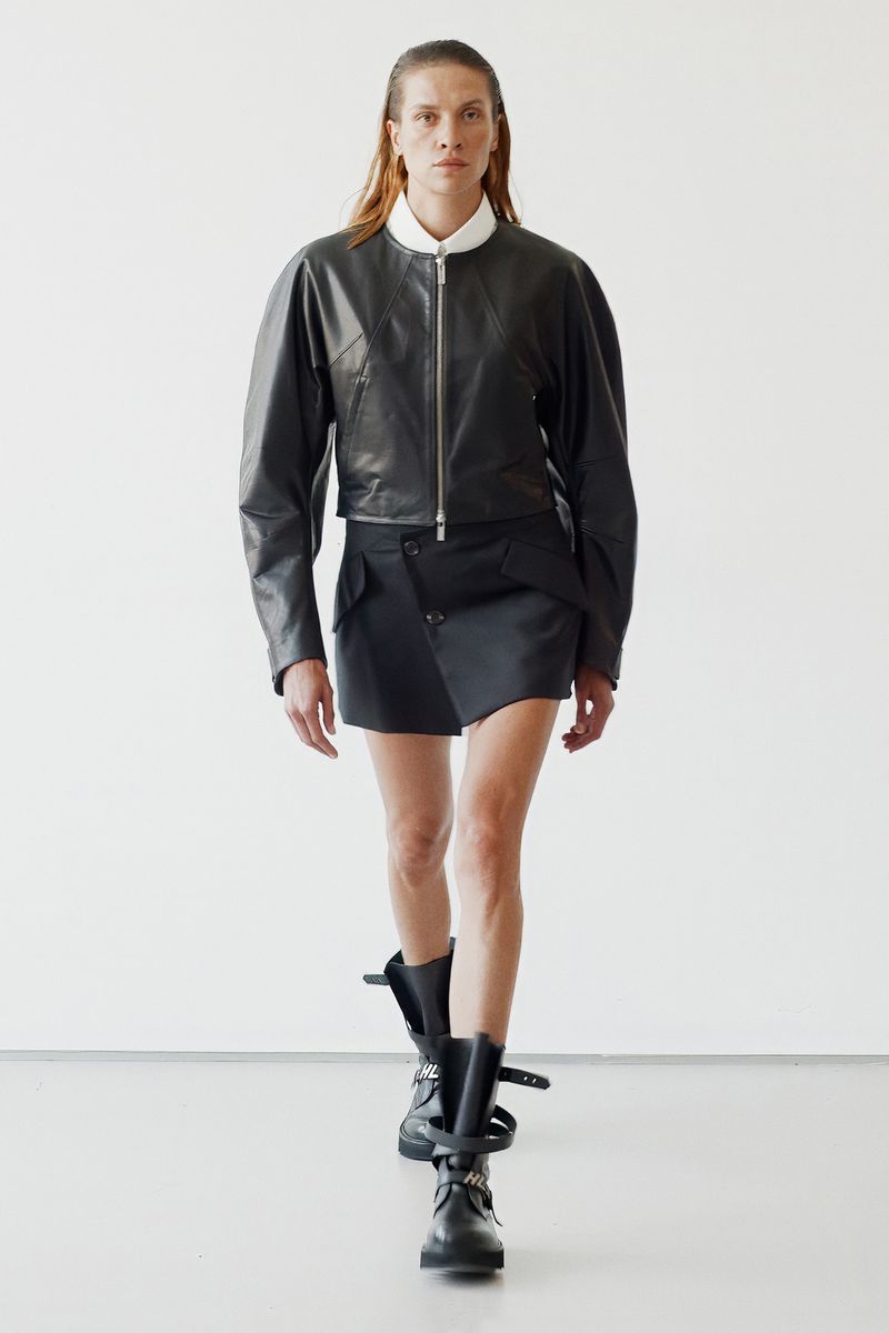 Model wearing a black leather jacket and asymmetric skirt against a plain white studio wall for Helmut Lang