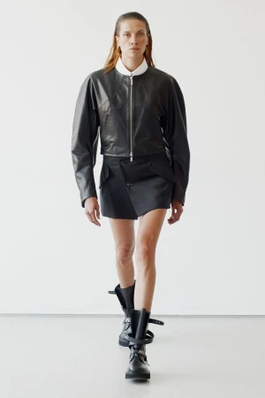 Model wearing a black leather jacket and asymmetric skirt against a plain white studio wall for Helmut Lang