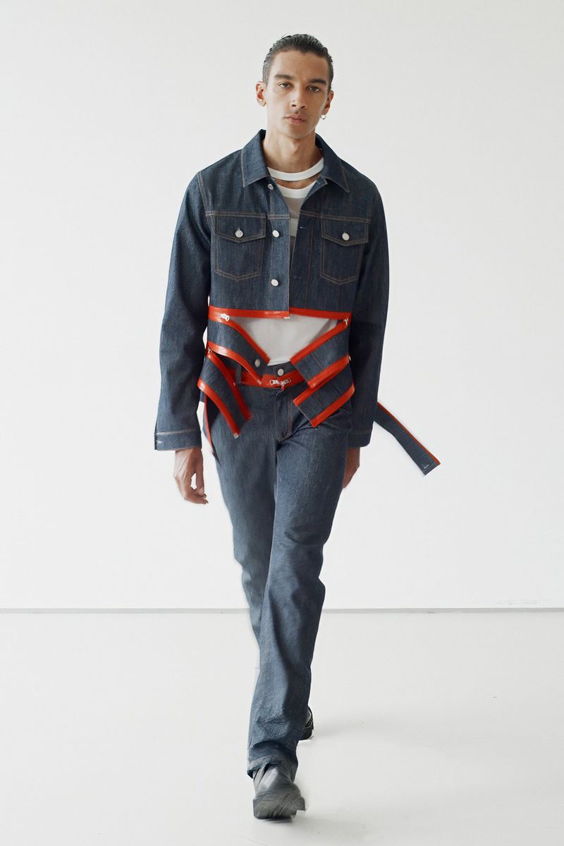 Fashion shot of a male model in a denim jacket and pants with a structural harness, posing in a white photo studio with a neutral expression.