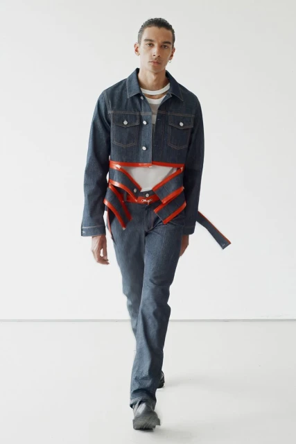 Fashion shot of a male model in a denim jacket and pants with a structural harness, posing in a white photo studio with a neutral expression.