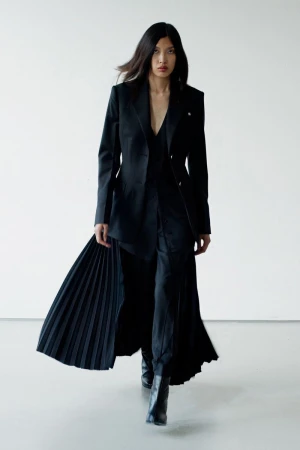 A woman with long, dark hair walks confidently in a black blazer, pleated skirt, pants, and ankle boots against a plain studio backdrop.