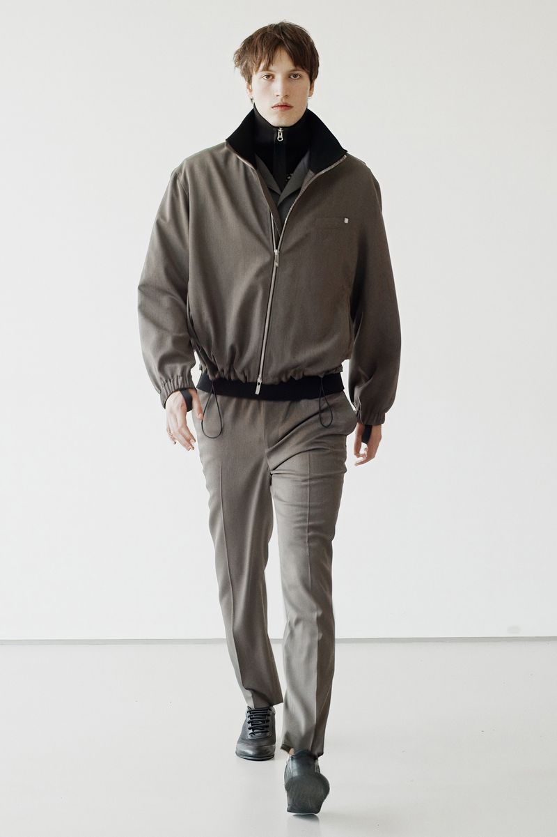 Male fashion model walks in earth-toned jacket and pants against a minimalist white backdrop, presenting contemporary style.