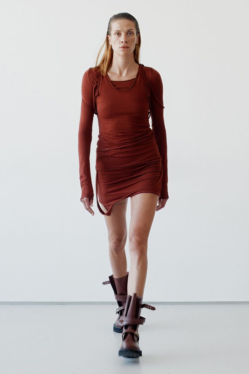 Fashionable adult woman in a brown mini dress and boots walks against a white wall in a studio, highlighting a contemporary fashion style.