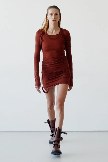 Fashionable adult woman in a brown mini dress and boots walks against a white wall in a studio, highlighting a contemporary fashion style.