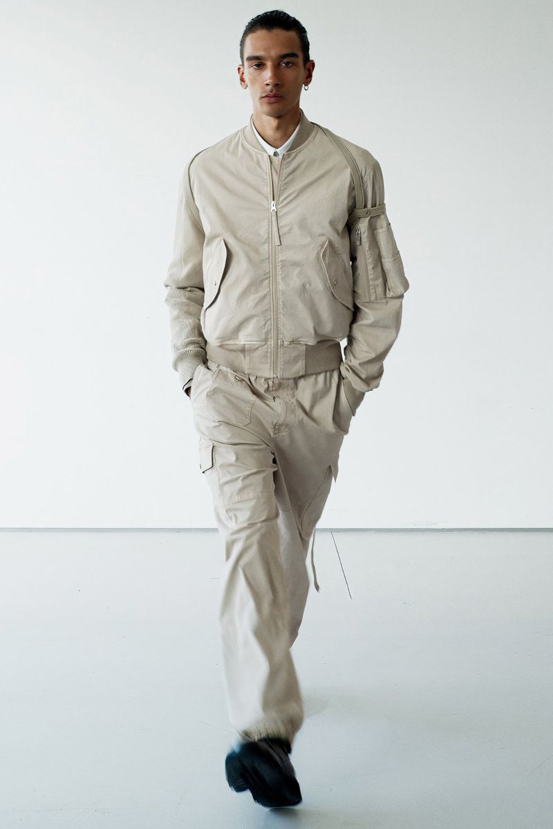 Adult male model in a relaxed pose wearing a beige bomber jacket and matching cargo pants, in a full-body shot against a white backdrop.