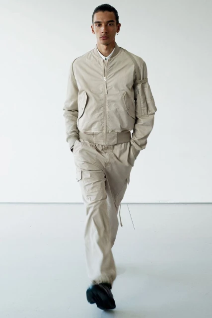 Adult male model in a relaxed pose wearing a beige bomber jacket and matching cargo pants, in a full-body shot against a white backdrop.