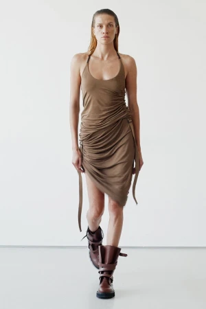 An adult female is walking and modeling a draped brown dress with brown, strapped boots against a plain white background in a studio shot.