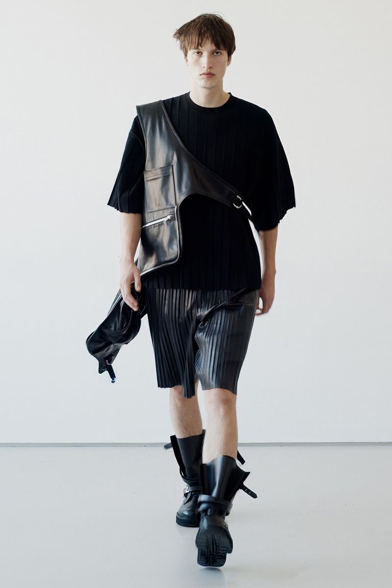 Male fashion model walks in black top, pleated shorts, leather vest, and boots against a white backdrop for a fashion editorial.