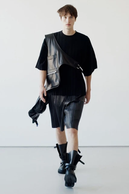 Male fashion model walks in black top, pleated shorts, leather vest, and boots against a white backdrop for a fashion editorial.