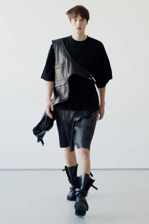 Male fashion model walks in black top, pleated shorts, leather vest, and boots against a white backdrop for a fashion editorial.