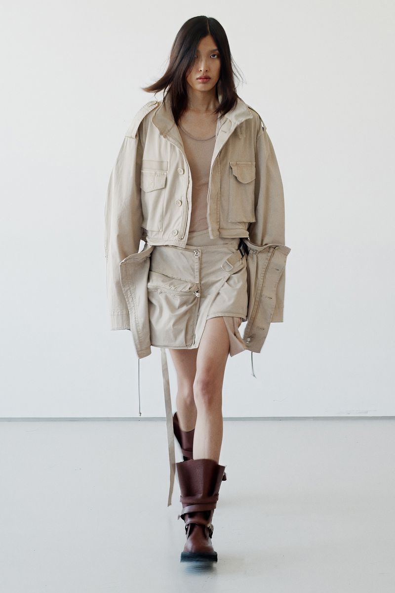 Fashion model presents tan jacket and skirt ensemble with brown boots in a minimalist studio setting during a runway fashion show.