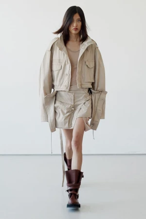Model walking forward wearing a beige utility jacket, skirt, and dark boots against a white wall.