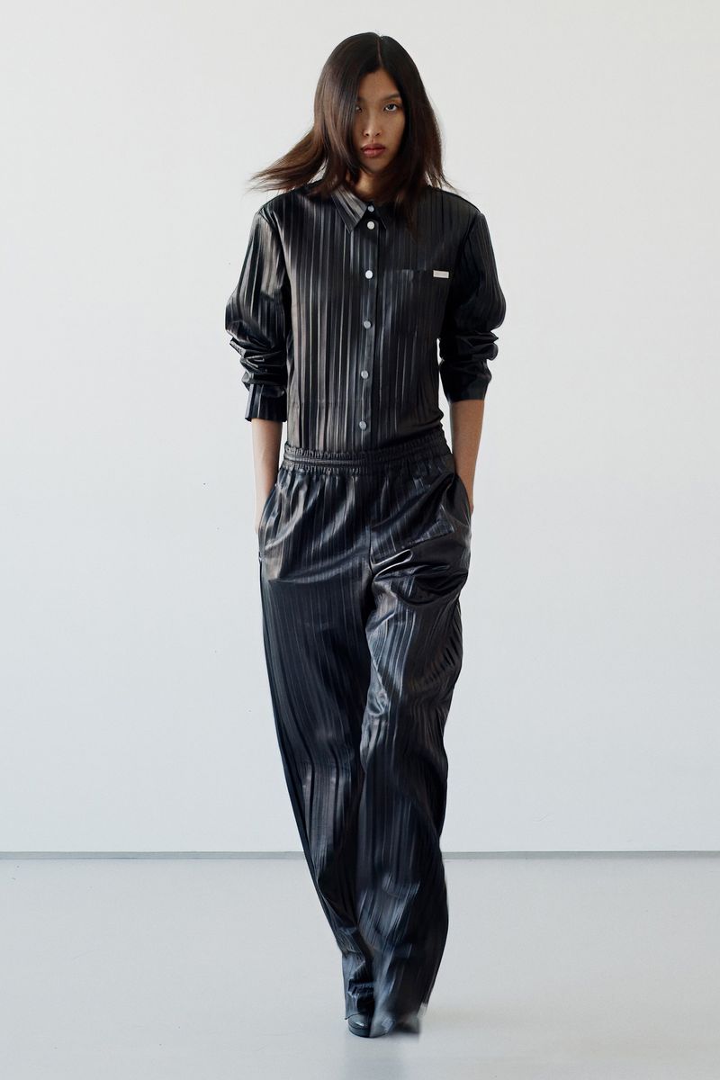 Fashion model poses in black outfit in a studio setting. She wears a shiny, vertically striped black shirt and matching wide-leg pants.