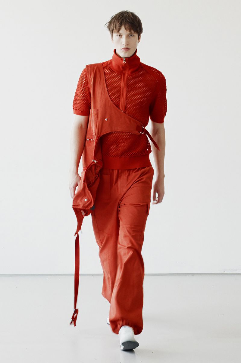A model is walking in a photo studio wearing a rust-colored knit shirt, cargo pants, and a matching harness with attached pouch and strap.