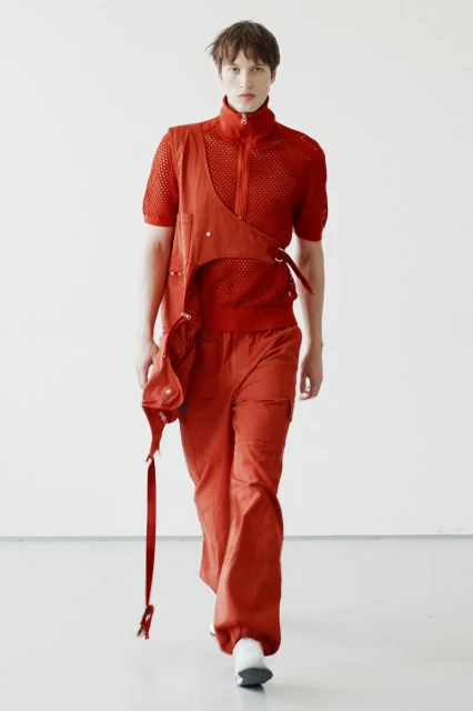 A model is walking in a photo studio wearing a rust-colored knit shirt, cargo pants, and a matching harness with attached pouch and strap.