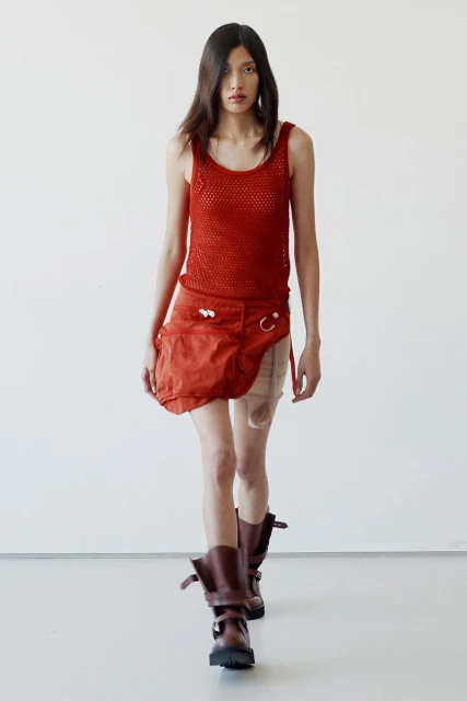 Fashion photo of a slender adult female model in a red tank top, distressed orange skirt, and chunky boots, walking on a white studio background.