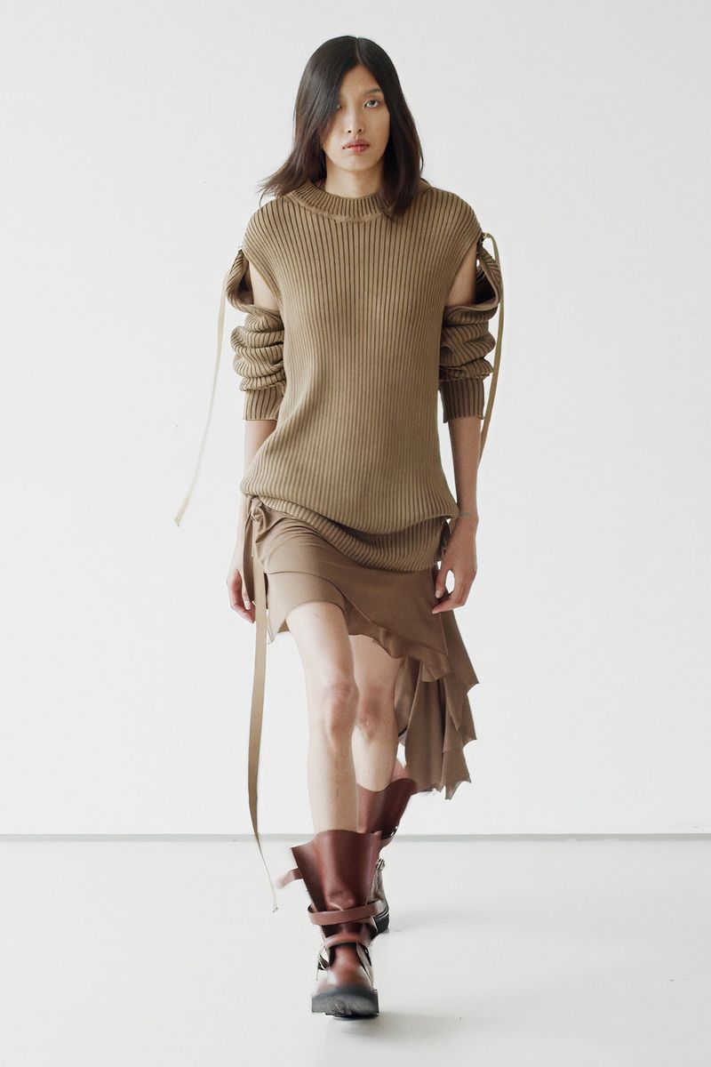 Fashion model walks runway in brown knit dress and platform boots against white background, showcasing modern apparel and footwear.