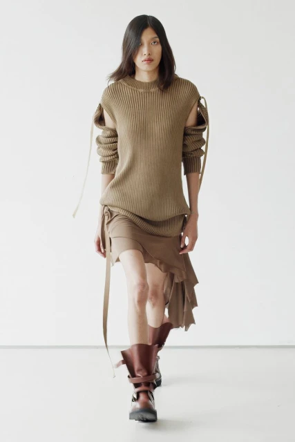 Fashion model walks runway in brown knit dress and platform boots against white background, showcasing modern apparel and footwear.