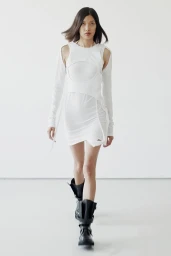 Model walking forward in a white cut-out dress and black boots against a plain white studio backdrop.