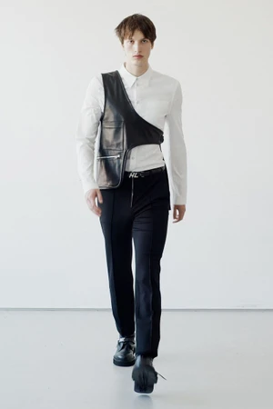 Model walking forward wearing a white shirt, black trousers, and an asymmetrical leather harness vest against a plain white wall