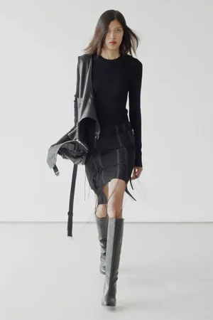 Model in black Helmut Lang outfit with knee-high boots and jacket walking against a plain white studio background.