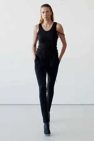Model in black tank top and matching trousers walking against a white studio background.