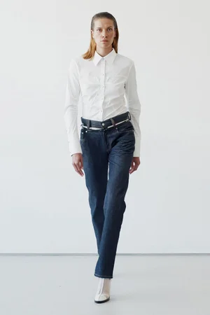 Model wearing a white button-down shirt and dark denim jeans standing against a plain white studio background.