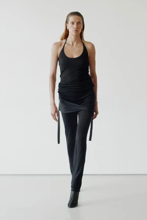Model in black draped top and slim trousers standing against a white seamless background
