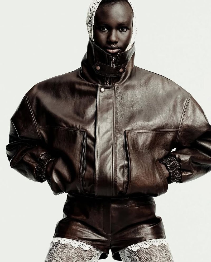 A dark-skinned model poses in a studio wearing a brown leather bomber jacket and shorts, and a white knit head covering.