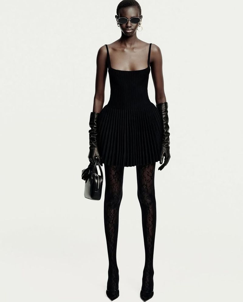 An adult woman with sunglasses poses in a short black dress, gloves, patterned tights, and heels against a bright white background.