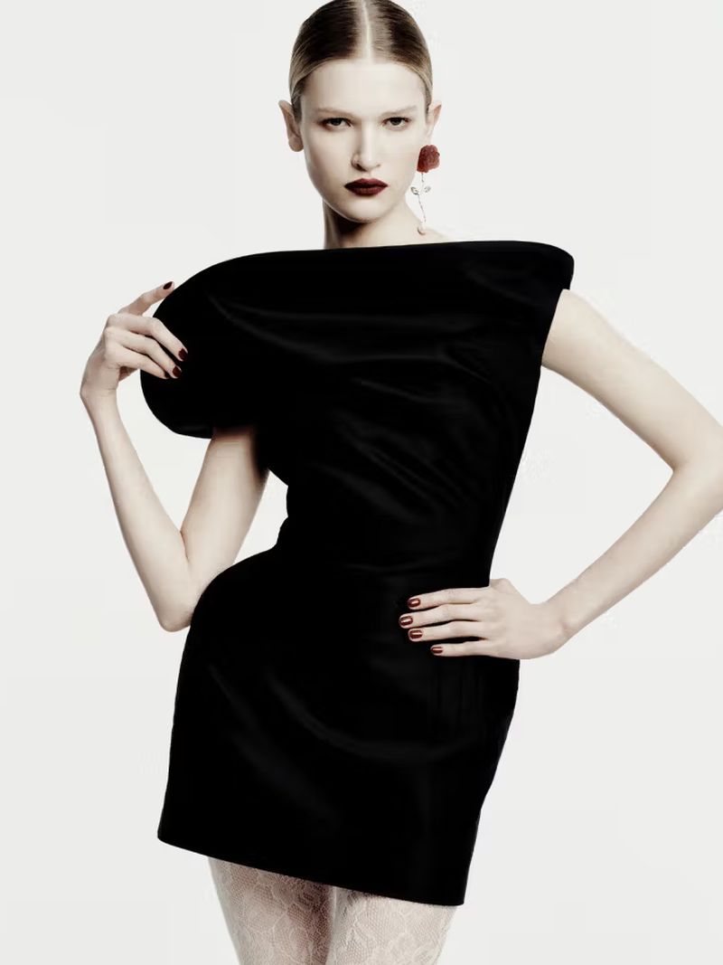 A young woman poses in a black mini dress with a dramatic shoulder in a studio fashion shot, with a stark white background.