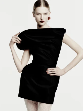 A young woman poses in a black mini dress with a dramatic shoulder in a studio fashion shot, with a stark white background.