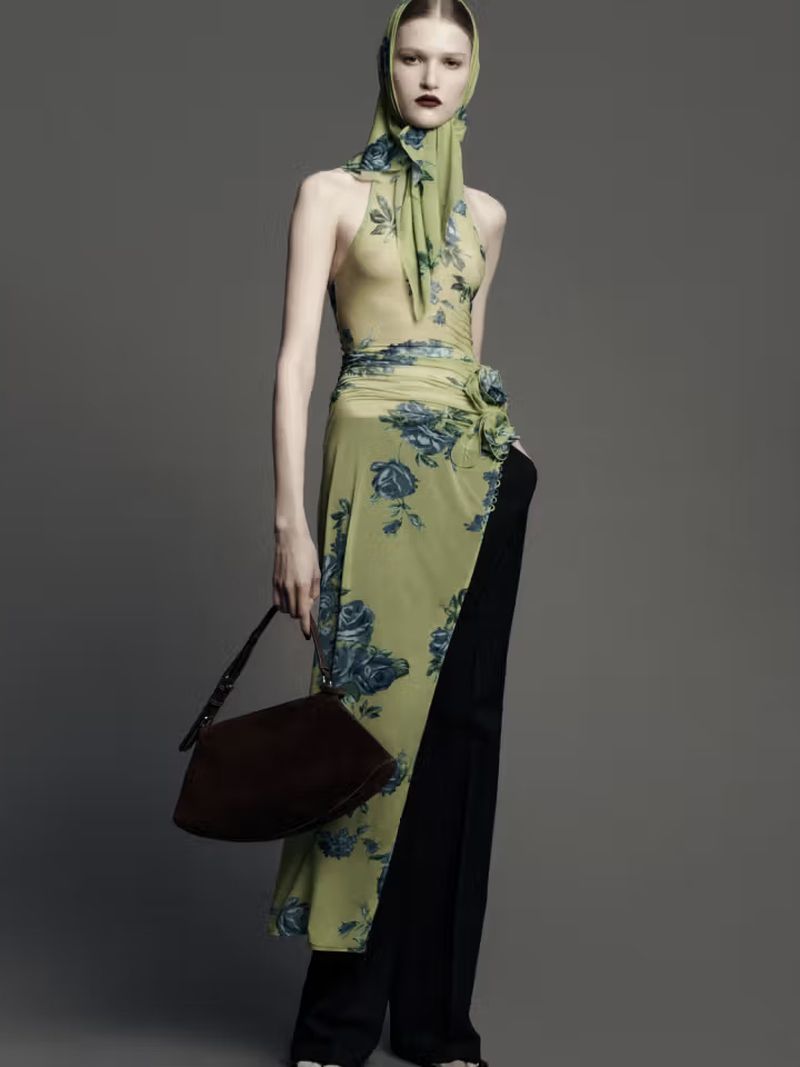 Fashion shot of a slender woman wearing a flowing floral dress and headscarf, holding a dark brown purse against a grey background.
